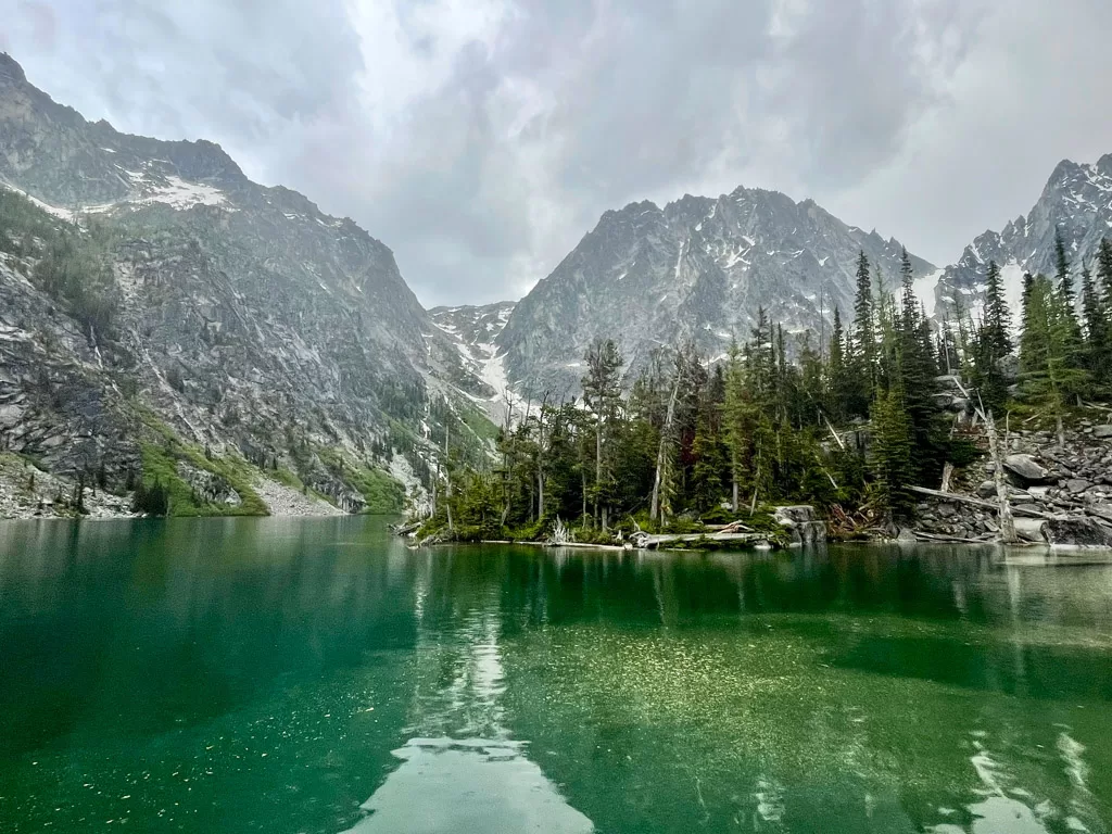A picture of Colchuck Lake a beautiful mountain lake with green water, big snowy mountains in the background and a beautiful forest USA bucket list adventure