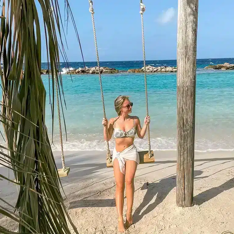 Blonde girl on swing in front of blue ocean at Mambo Beach Boulevard with a decorative element of palm leaves in the foreground