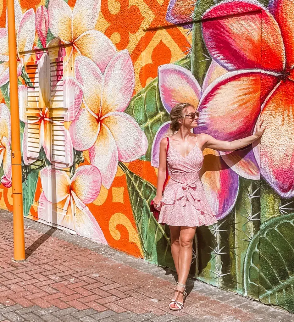 Beautiful mural made of orange, pink and many other colors with flowers and patterns and author standing in a dress in front of it 