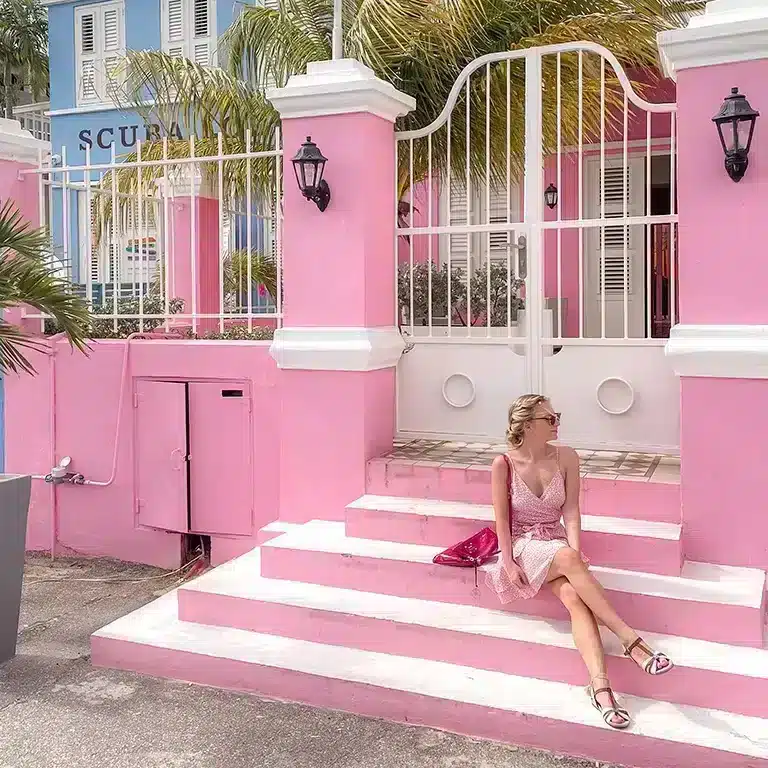 Blonde girl sitting on the steps of a pink building in willemstad