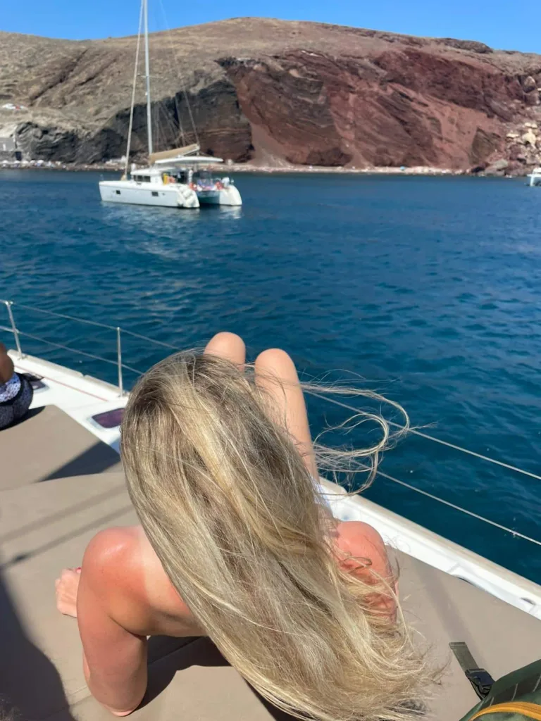 blonde girl lying on a boat relaxing in front of red beach santorini 