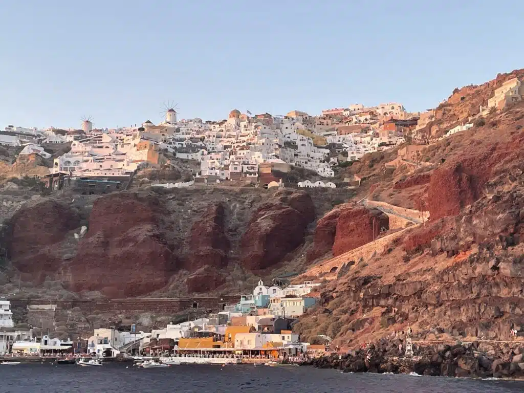 picture of famous ammoudi bay in santorini with impressive red cliffs and town with white houses on top of it taken from boat