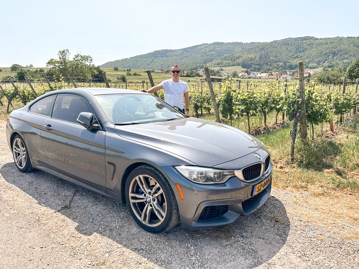 the authors husband on a road trip with his car in front of the vineyards in the Alsace, a spot totally worth visiting