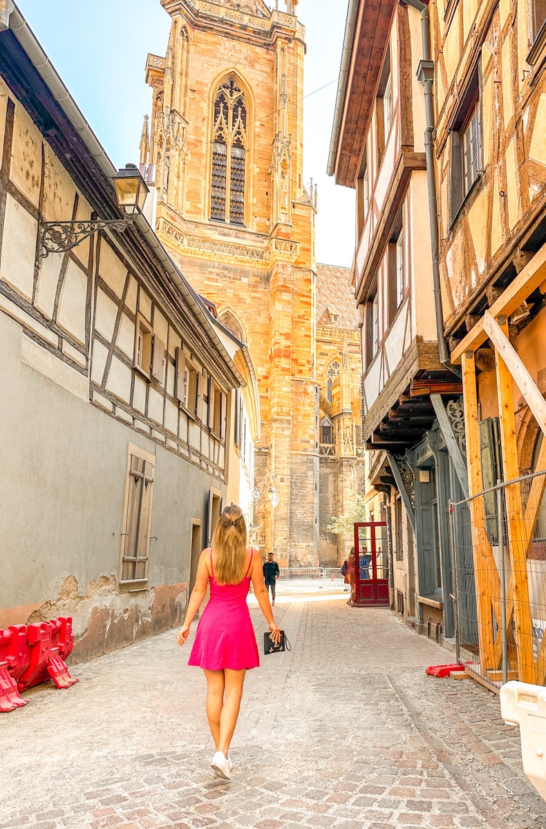 picture of the author in a pink dress in colmar walking down a street toward a cathedral