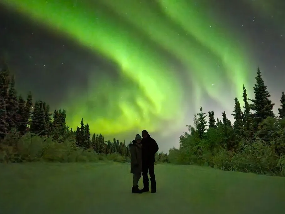 the authors friend and her husband in front of the northern lights in alaska on a winter road trip 