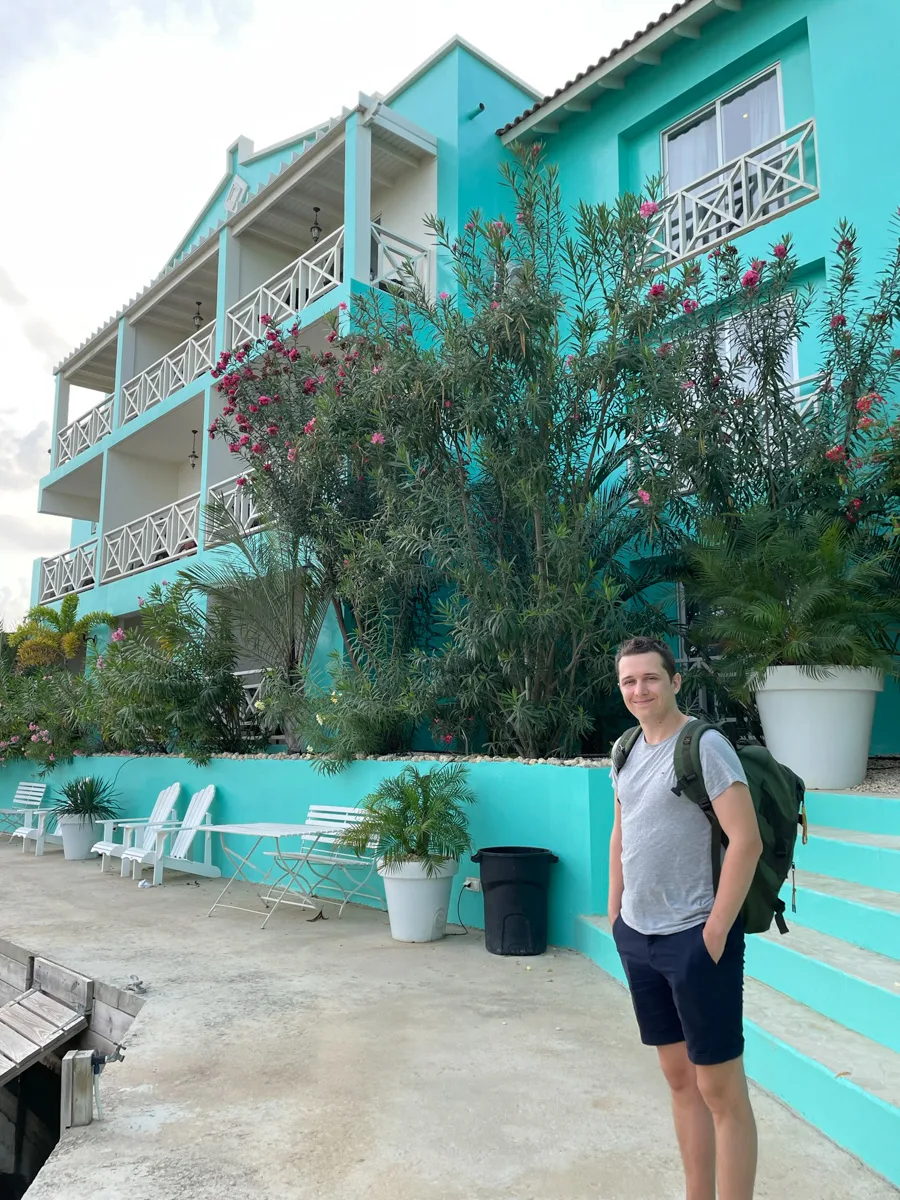 A male companion stands in front of a multi-level, pastel-colored building with a spacious outdoor seating area in Bonaire, capturing the island's relaxed atmosphere.