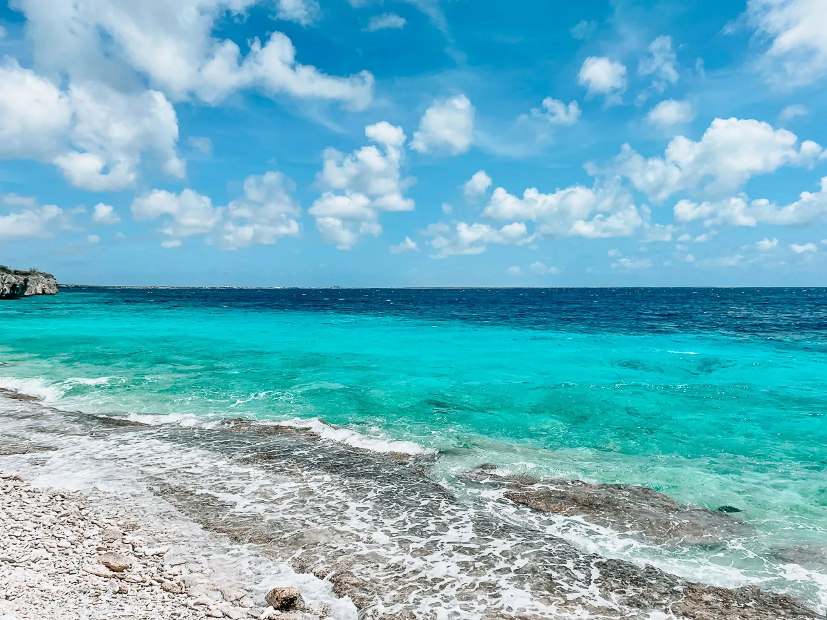 Image of the crystalline turquoise waters of Bonaire. The sea is calm with gentle waves lapping onto a pebbly shore. Clouds scatter across the bright blue sky, reflecting the sun's radiance.