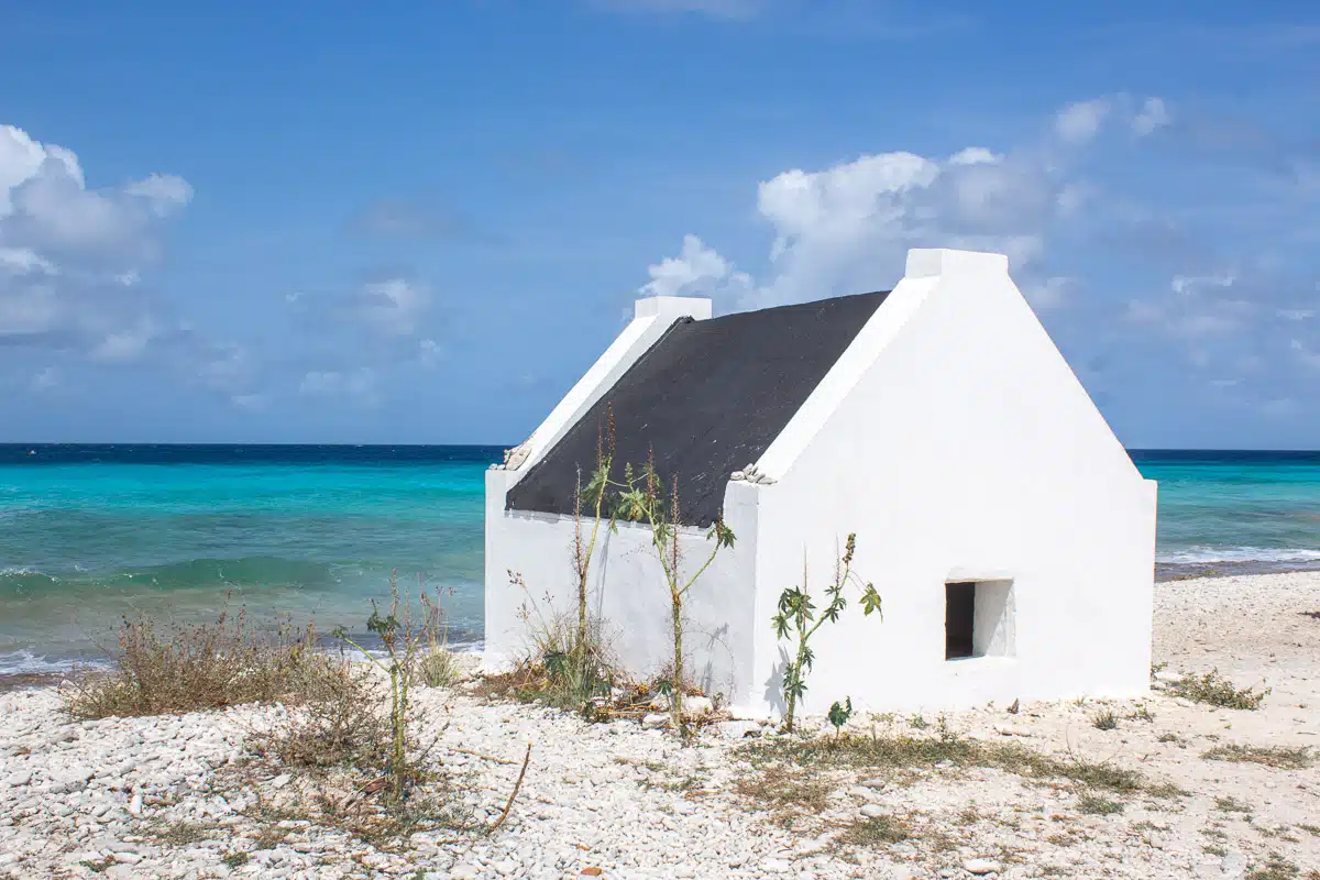 A simple, white, traditional stone hut stands alone on a Bonaire beach, with the Caribbean Sea stretching out behind it under a sky partially filled with cumulus clouds.