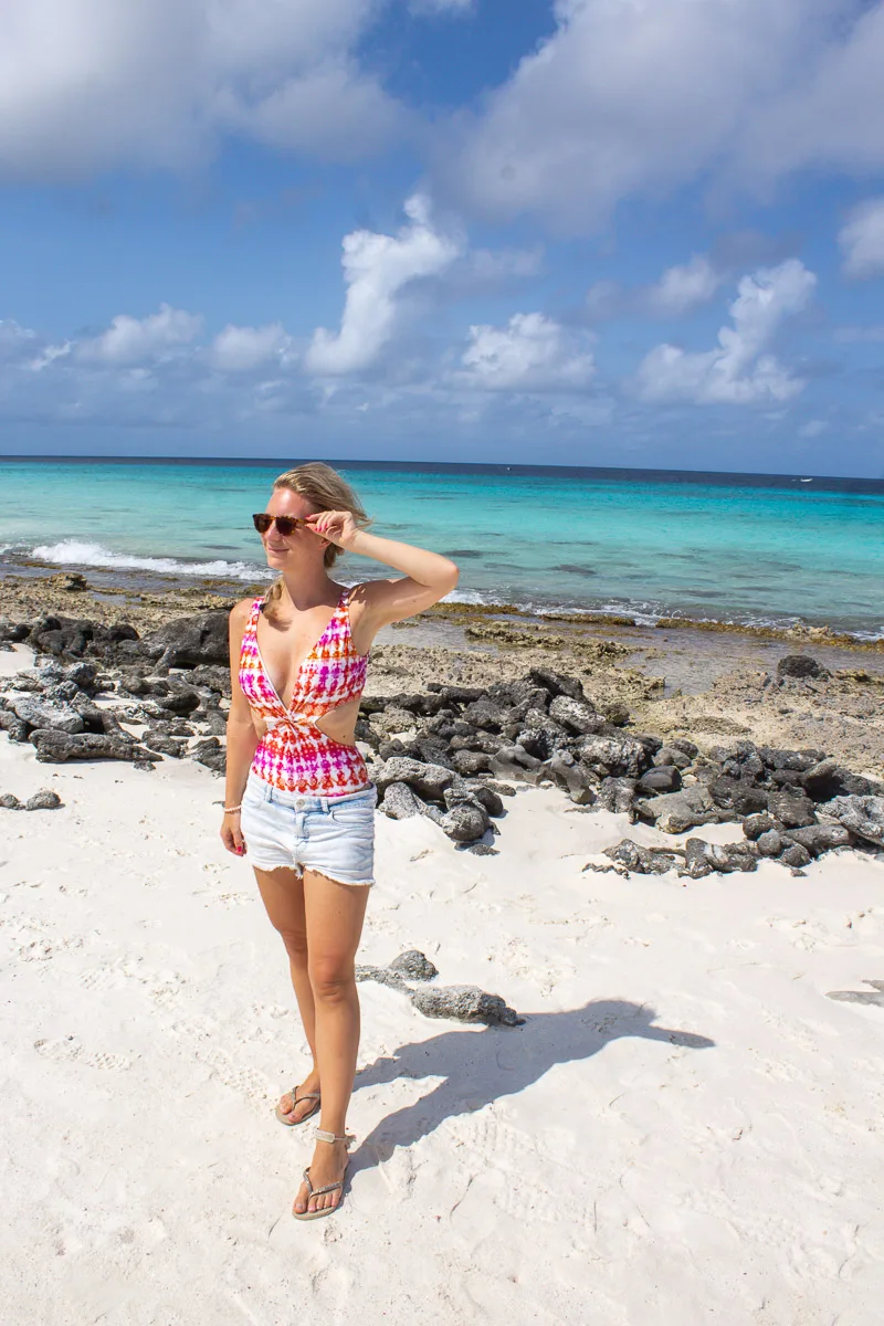 The author stands on a rocky beach in Bonaire. She's dressed in a red and white halter top and denim shorts, with sunglasses on and the clear blue sea behind her.