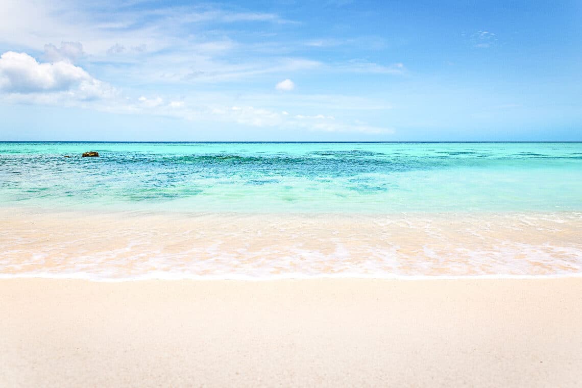 Pristine sandy beach meeting the crystal-clear turquoise waters of a calm sea, under a beautiful sky with soft clouds, creating a serene tropical scene.