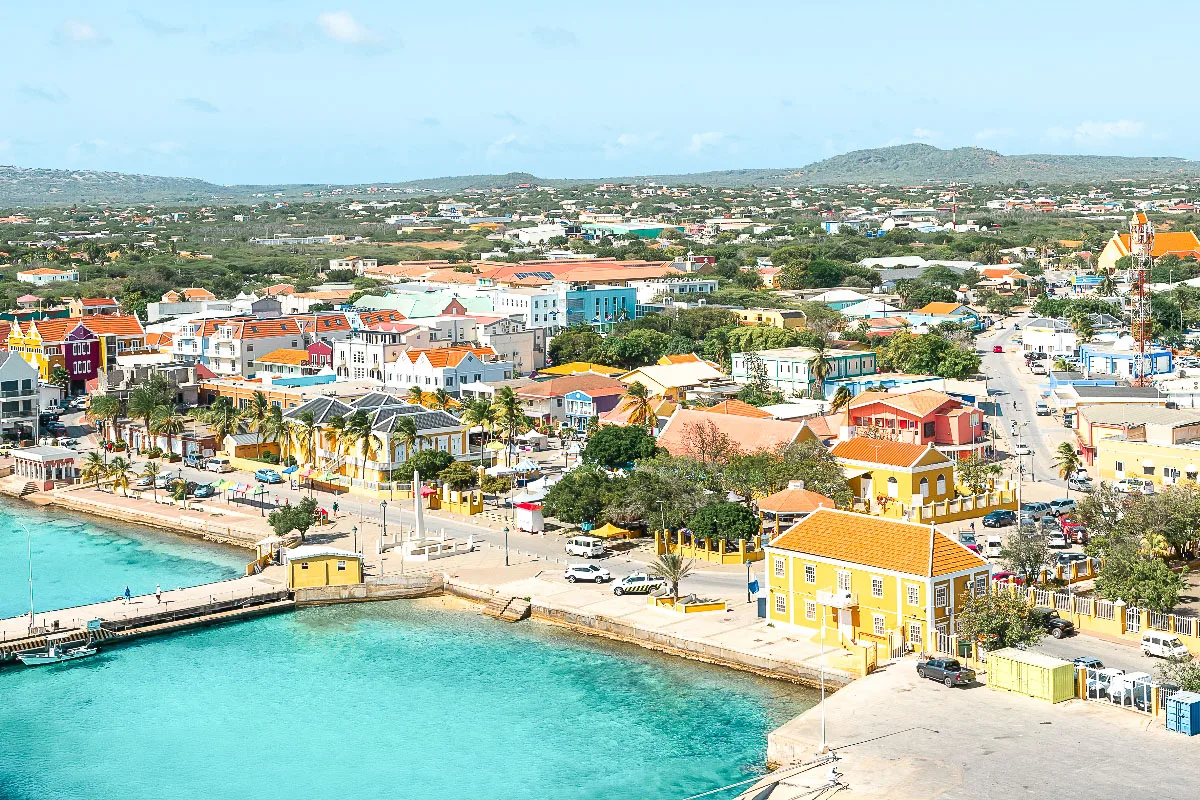 Picture of the Bonaire Cruise Port and Kralendijk in the background. A panoramic view of a colorful coastal town with a waterfront promenade, featuring a variety of brightly painted buildings and a clear blue sky overhead.