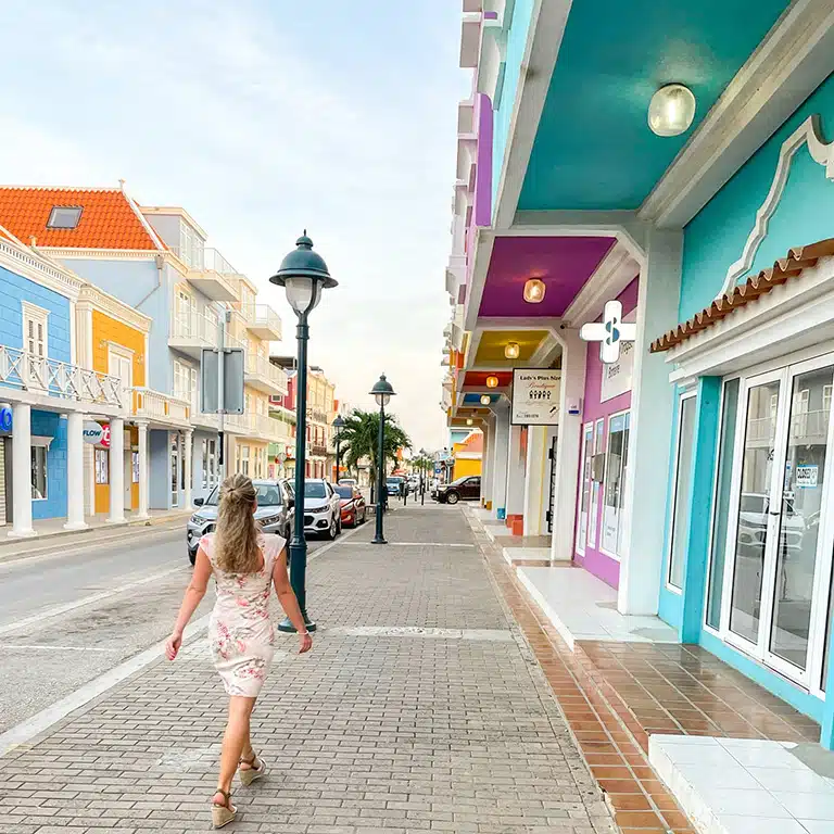 A picturesque street scene in Kralendijk, Bonaire. Colorful buildings with European architectural influences line the sidewalk. The woman, likely the author, walks away from the camera, adding a sense of exploration.
