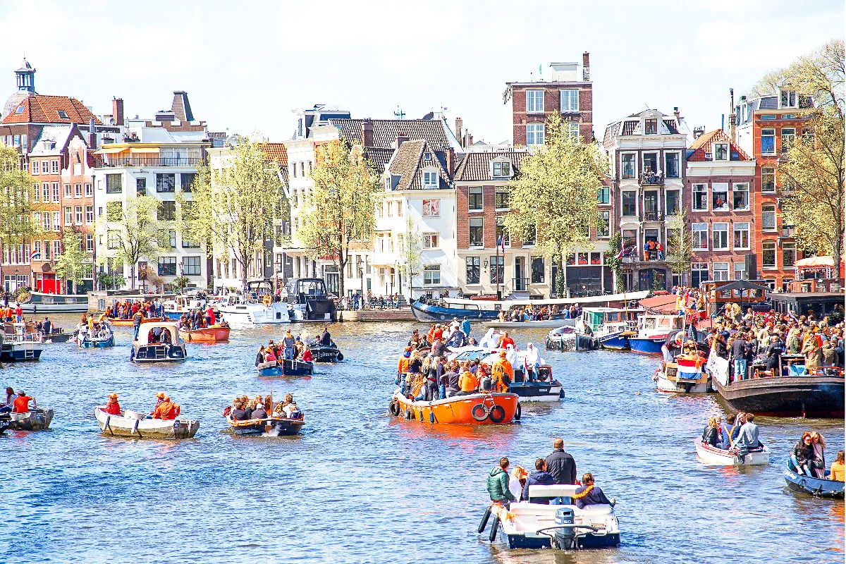 Image of a bustling canal in Amsterdam, lined with historic Dutch buildings and filled with boats. People are celebrating on boats, many dressed in bright orange, possibly during a festival or national holiday.