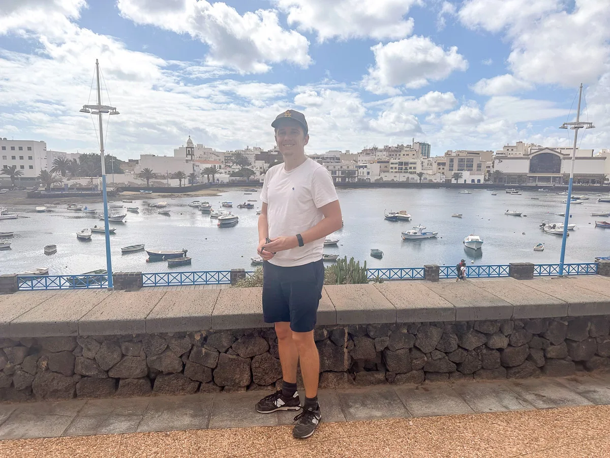 my husband in a white t-shirt and baseball cap stands smiling in front of the scenic Charco de San Ginés lagoon, filled with small boats, in Arrecife, Lanzarote, under a partly cloudy sky.