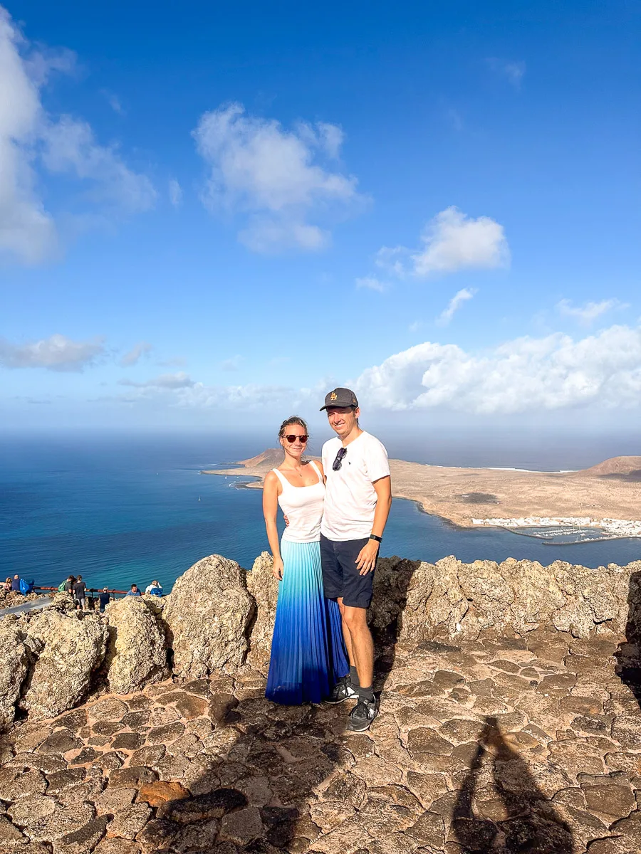 us standing atop the rocky viewpoint at Mirador del Rio, Lanzarote, with panoramic views of the sea and La Graciosa island under a bright blue sky with fluffy clouds."