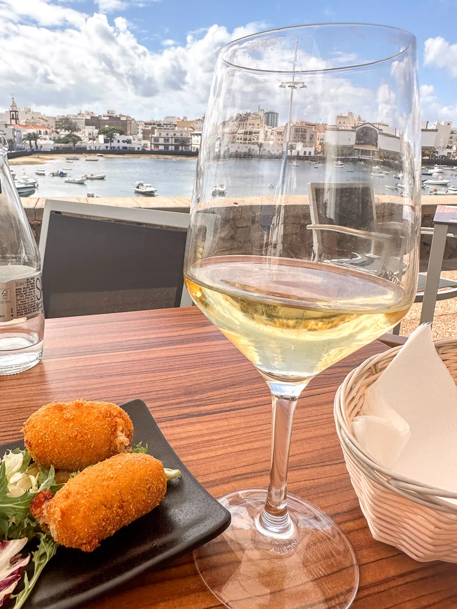 A close-up view of a glass of white wine and two croquettes on a plate, with a blurred background showcasing the Charco de San Ginés lagoon and the Arrecife skyline."