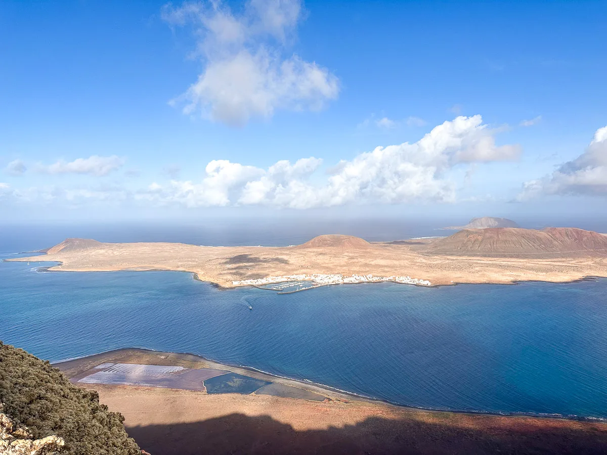 "An aerial perspective from Mirador del Rio, overlooking the azure waters surrounding La Graciosa island, with the small village and sandy terrain visible under a sky dotted with clouds."