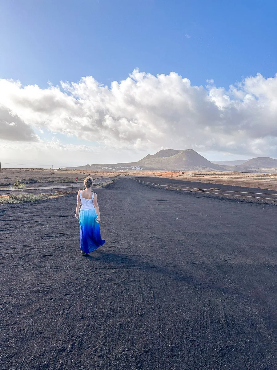 the author walks on a black volcanic ash road in Lanzarote, looking towards distant mountains under a vast sky with scattered clouds, embodying a sense of exploration."