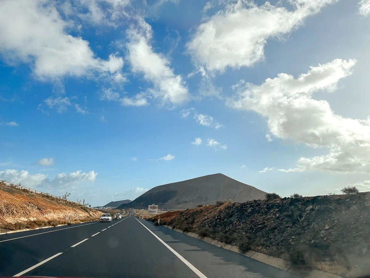 "A scenic view from Lanzarote, showcasing the rugged volcanic landscape with a view of the coastline and the road leading towards distant mountains under a partly cloudy sky."