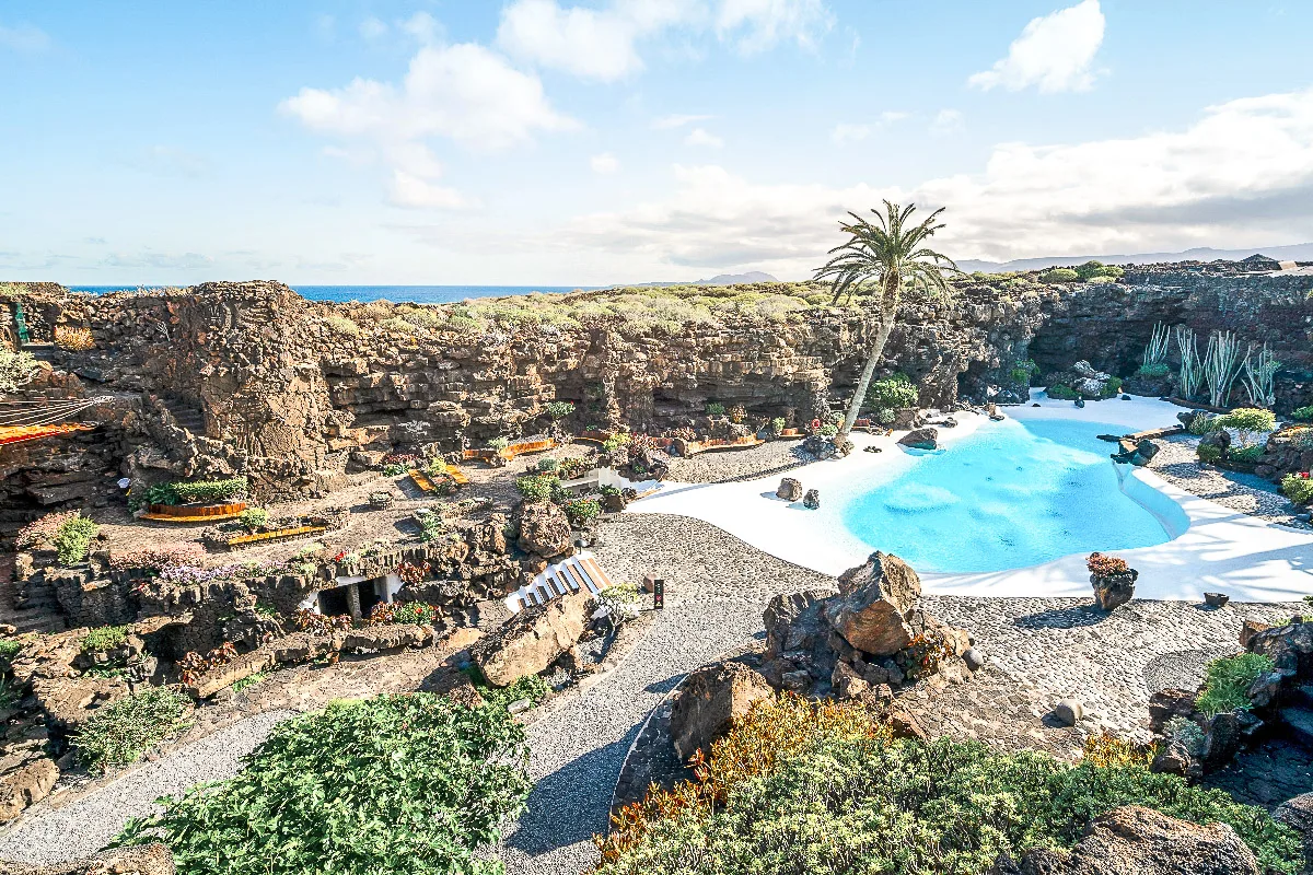 The luxurious and exotic pool area of Jameos del Agua, Lanzarote, nestled within a volcanic rock formation, with lush vegetation and a single palm tree, under a clear blue sky."