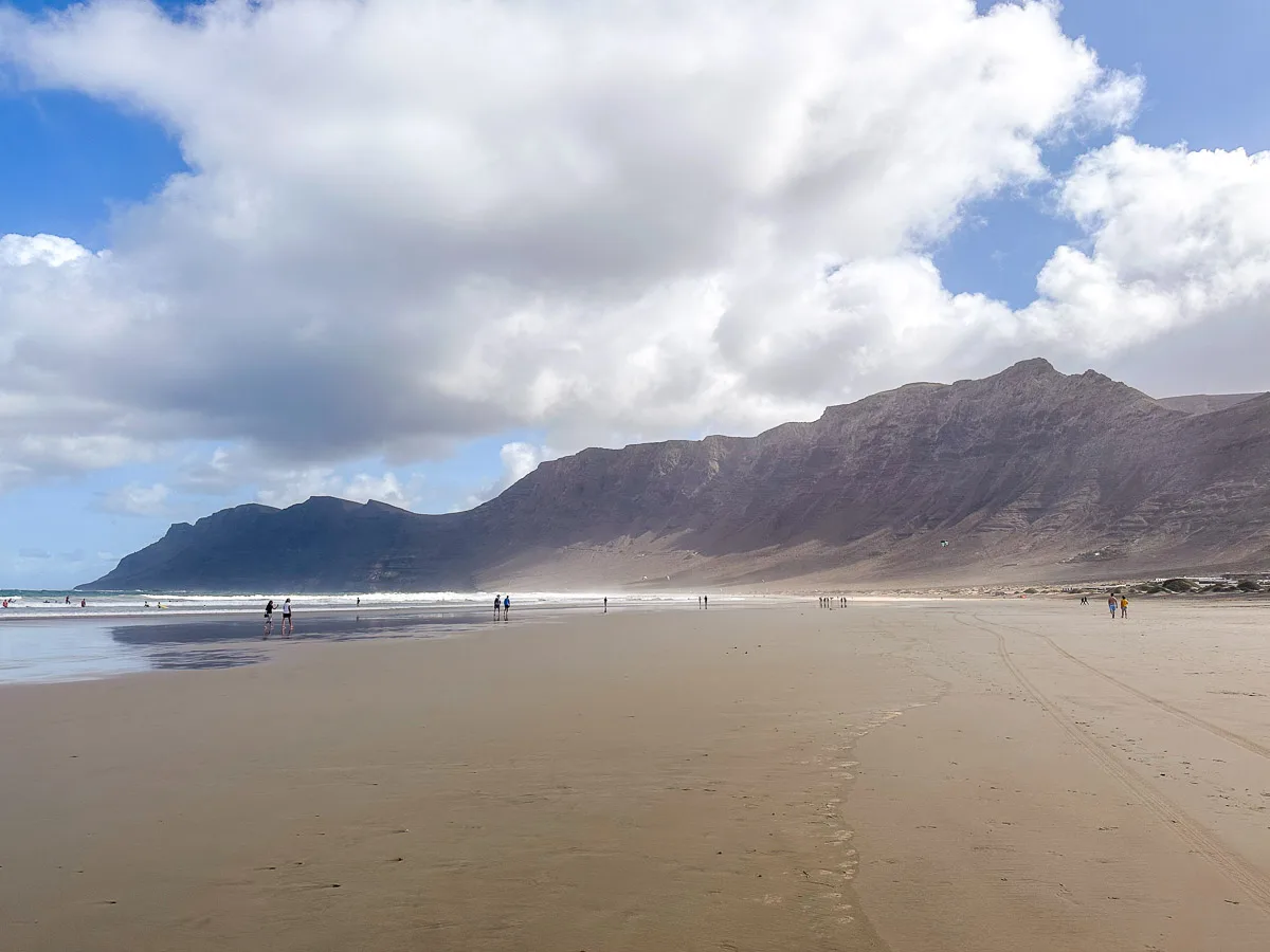 A serene view of Famara Beach in Lanzarote with expansive sandy shores and the majestic Risco de Famara cliffs under a partly cloudy sky, with visitors enjoying the vast beach