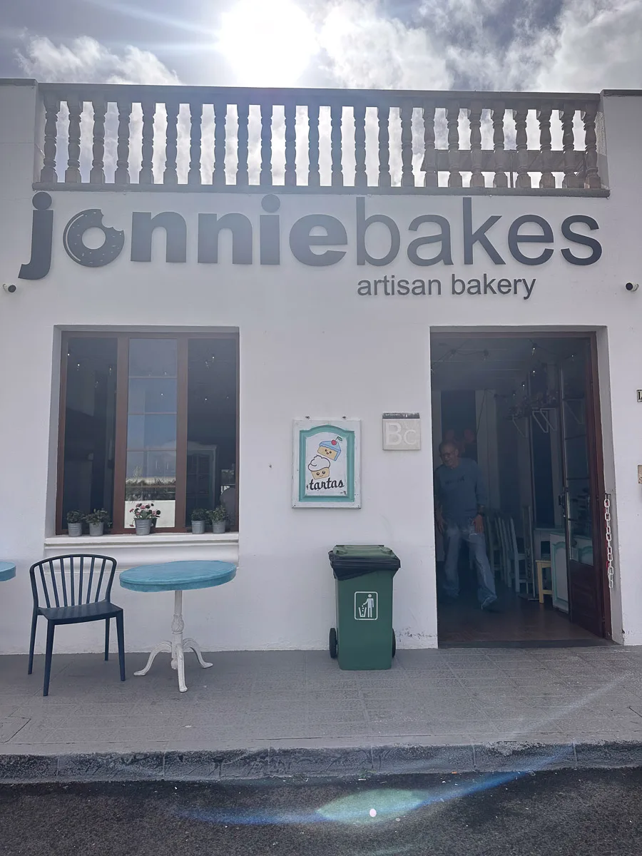 The front of Jonnie Bakes artisan bakery in Lanzarote, featuring a clean, white façade with bold lettering, a turquoise table, and a green waste bin, under a bright sky