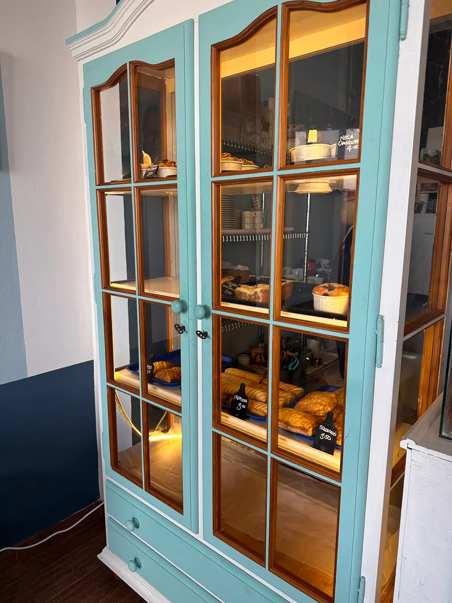 A display cabinet with a turquoise frame at Jonnie Bakes bakery, showcasing an appetizing array of baked goods like croissants and cakes, invitingly illuminated from within