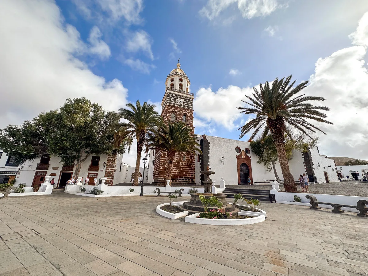 The historic Teguise Church in Lanzarote, framed by palm trees and a clear sky, with its iconic bell tower and traditional white-washed walls, as visitors explore the surrounding square