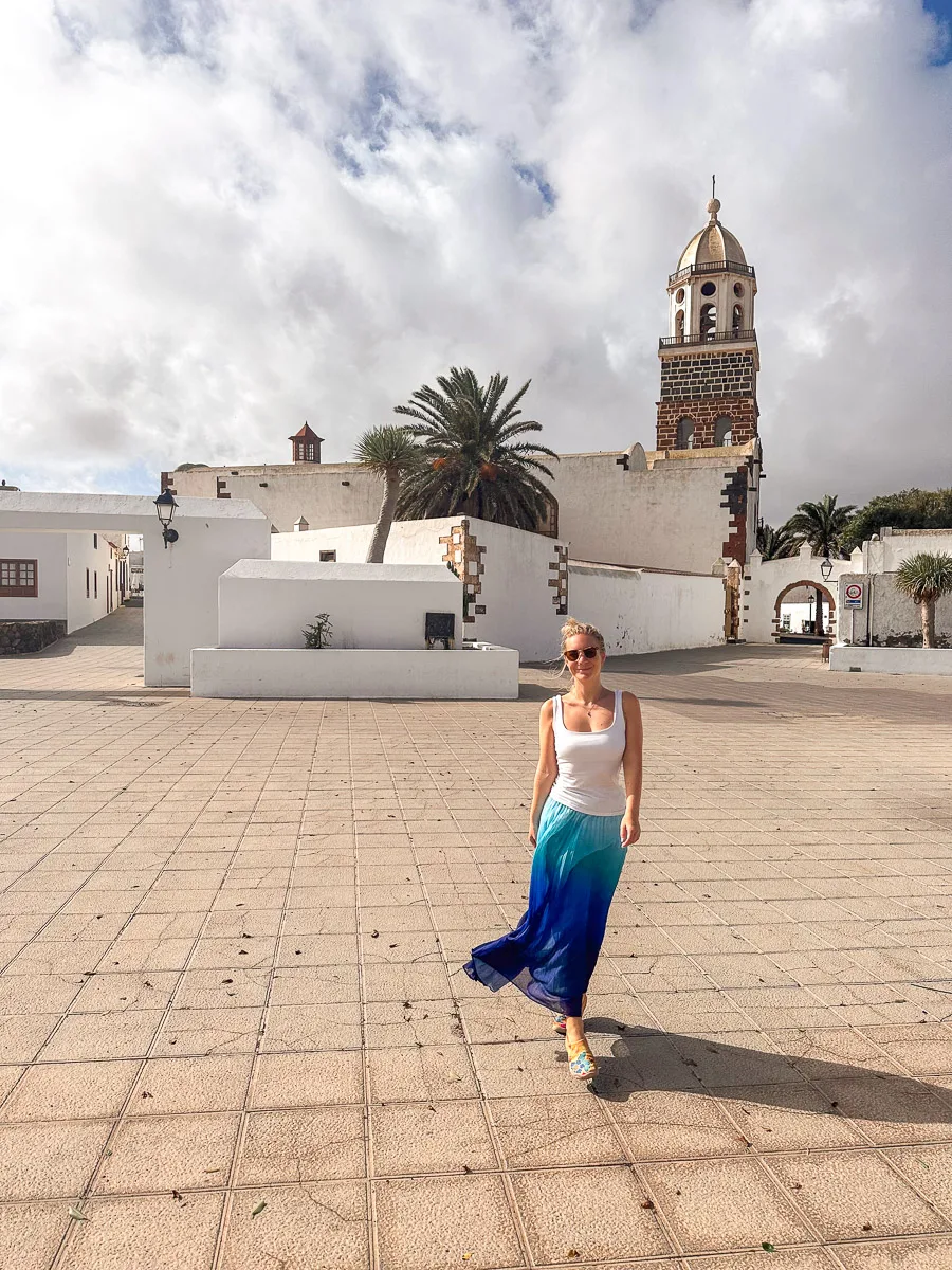 The author in a white tank top and blue skirt playfully poses in the plaza of Teguise Church in Lanzarote, with the church's bell tower and palm trees in the background