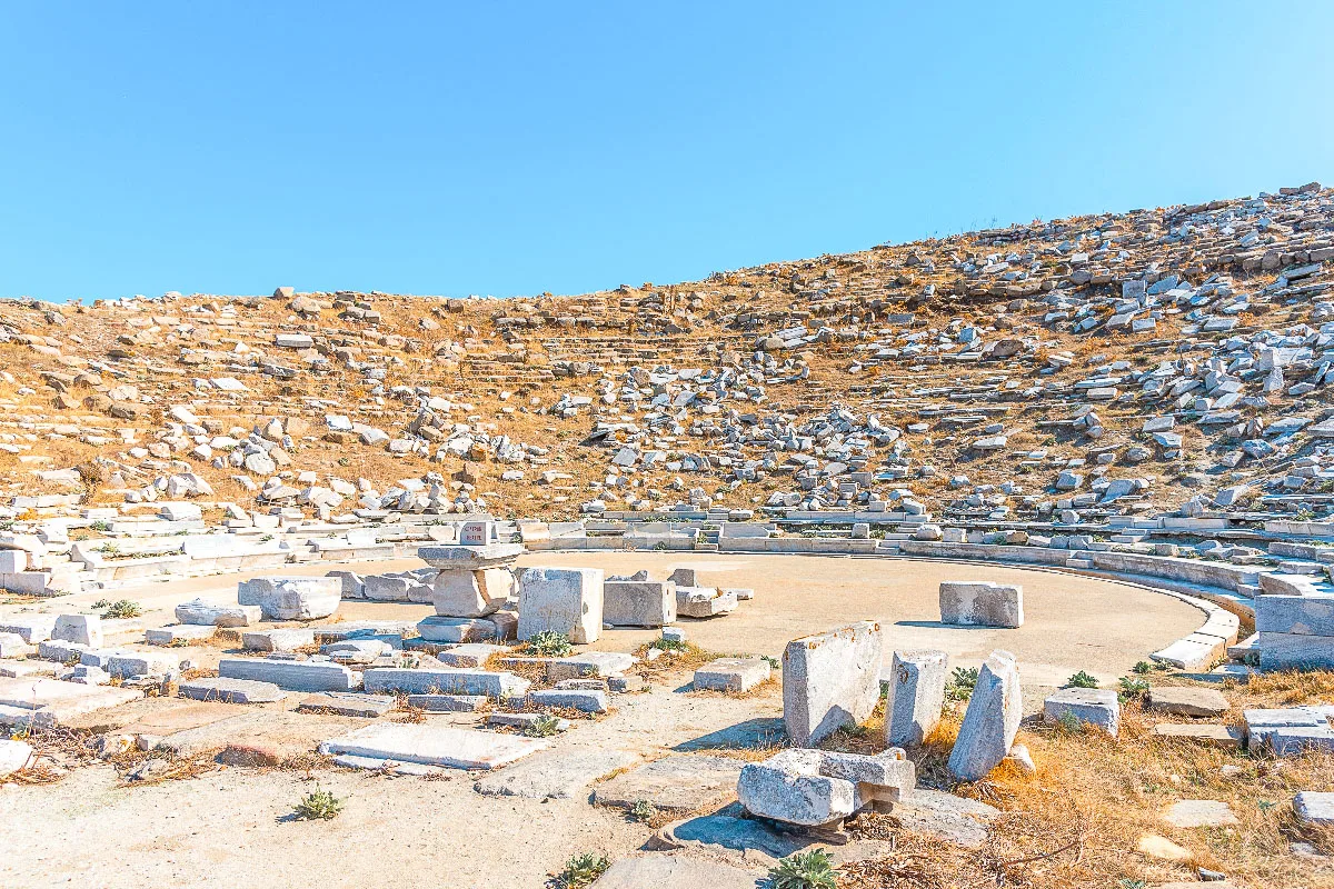 The ancient ruins of a circular, stone-built assembly place, possibly an amphitheater. Sun-bleached stones and fragments are scattered across the ground, all under the vast expanse of a clear blue sky.