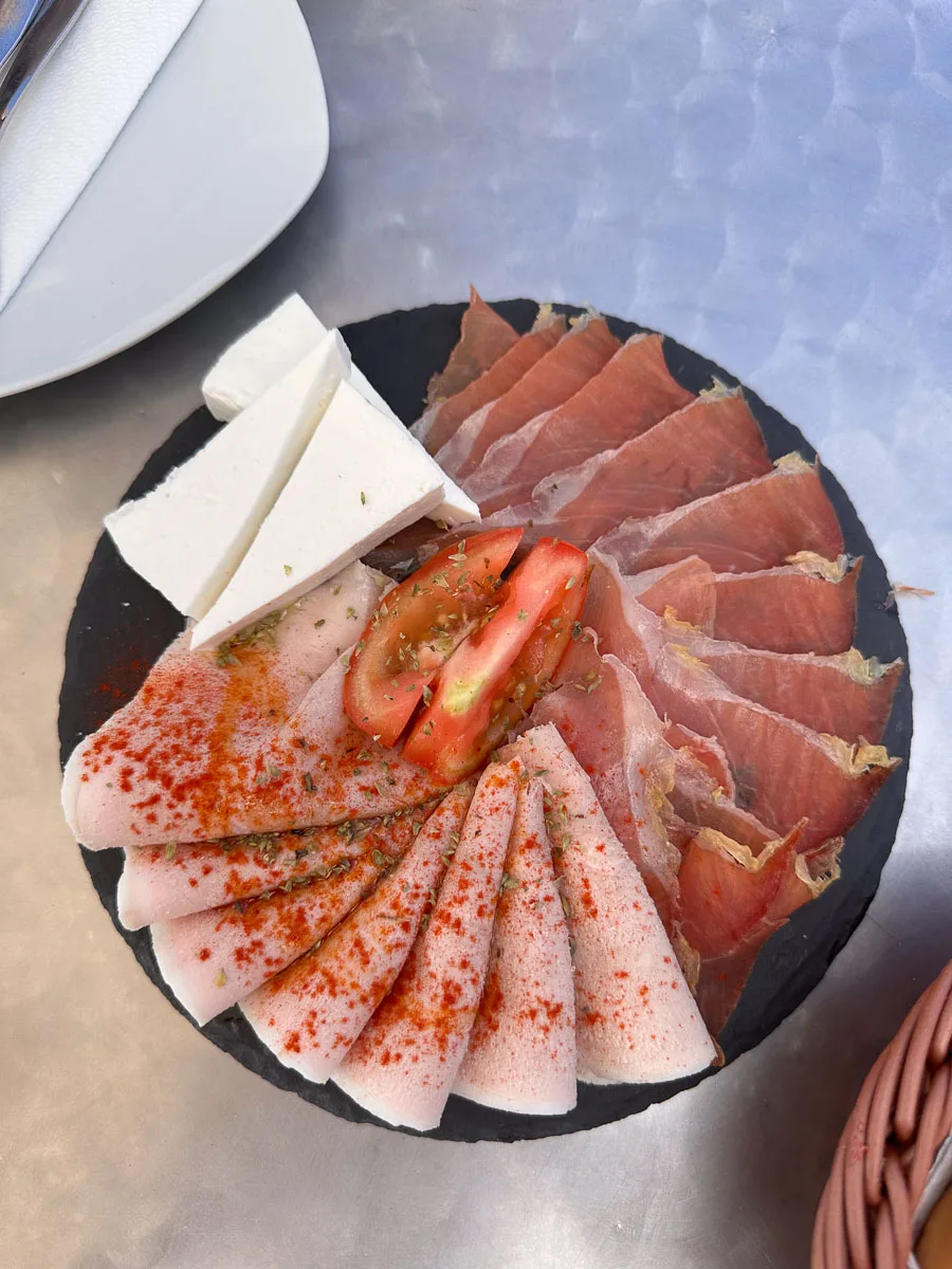 A plate of traditional Canarian cuisine in Tenerife, featuring thinly sliced cured ham, white cheese, and tomato dressed with herbs and a sprinkle of red paprika on a black slate serving board, showcasing the simple yet flavorful local fare.