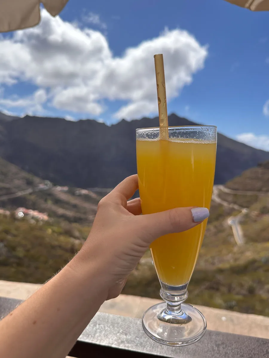A person's hand holding a glass of fresh orange juice with a bamboo straw, set against the backdrop of Tenerife's mountainous landscape under a sunny sky, encapsulating a moment of relaxation and refreshment during a day of exploration.