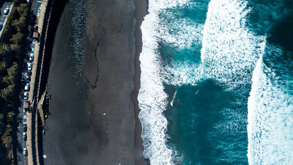 A top-down view of Tenerife's coastline showing the white foam of the waves crashing against the black volcanic sand beach, the contrasting colors and textures beautifully illustrate the dynamic and unspoiled nature of the island's beaches.