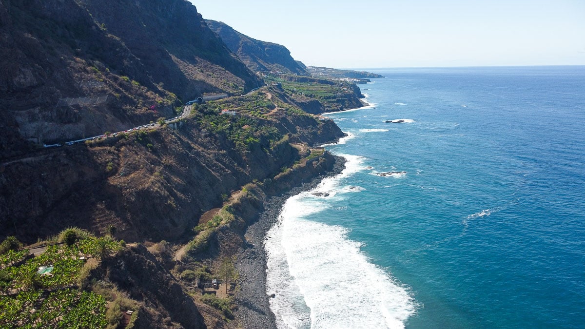 A coastal aerial shot showing a winding road that hugs the steep cliffs of Tenerife's shoreline, revealing the island's dramatic topography and the vast Atlantic Ocean extending into the horizon, embodying the adventurous spirit of the island.