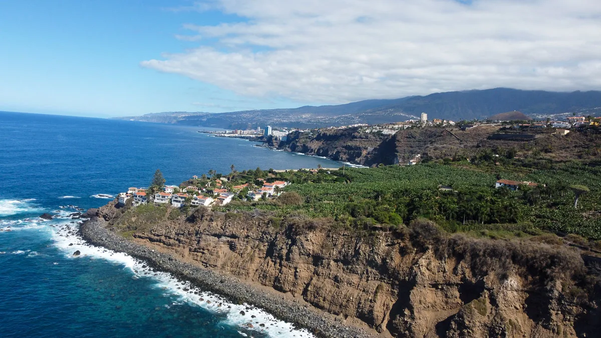 An aerial shot of the rugged coastline of Tenerife, with waves crashing against the cliffs. Residential areas with scattered houses are surrounded by banana plantations, showcasing the island's agricultural side amidst the dramatic coastal scenery, with the sprawling cityscape visible in the distance.