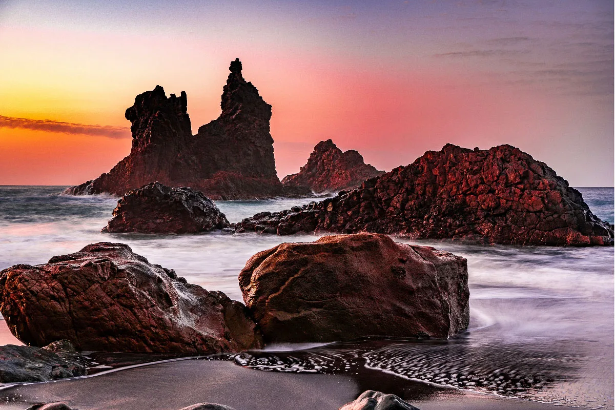 A stunning landscape shot capturing the rugged rock formations on a Tenerife beach at sunset, with the sky painted in hues of pink and orange, the long exposure creates a silky effect on the water, enhancing the scene's serene atmosphere.