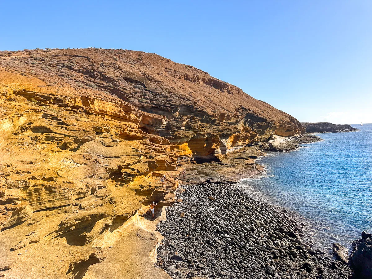 A secluded black pebble beach in Tenerife nestled at the base of a steep, rocky cliffside. The clear blue waters gently lap against the shore, and the layers of sedimentary rock in the cliff display a rich, earthy palette of colors, contributing to the serene and untouched feel of this picturesque coastal spot.