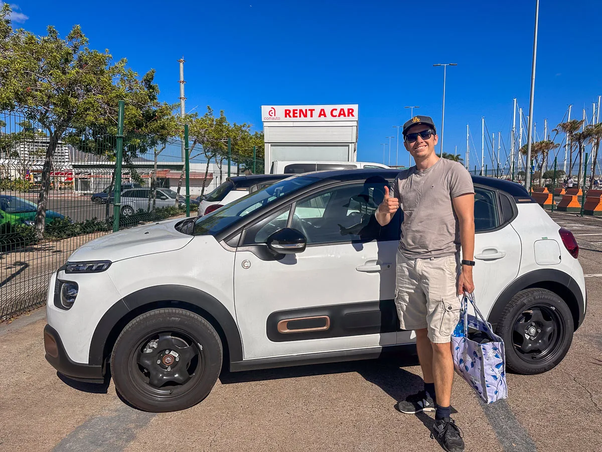 The author and her husband are standing in a car rental parking lot in Tenerife, ready for an adventure. They're beside a stylish white compact SUV, with the author's husband giving a thumbs-up. The marina in the background hints at the coastal explorations that await them, with a clear blue sky above and lush palm trees swaying gently.