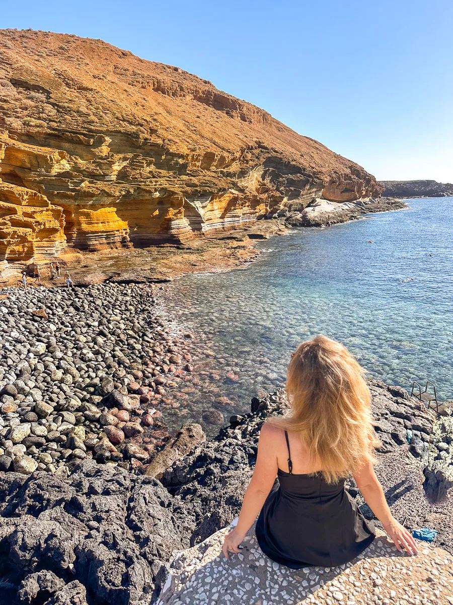 The author is seated on a mosaic-tiled surface, gazing out at the tranquil waters of a secluded cove in Tenerife. The contrasting black volcanic rocks border the clear sea, and the rugged cliffs provide a natural backdrop that highlights the island's volcanic origins and serene beauty.