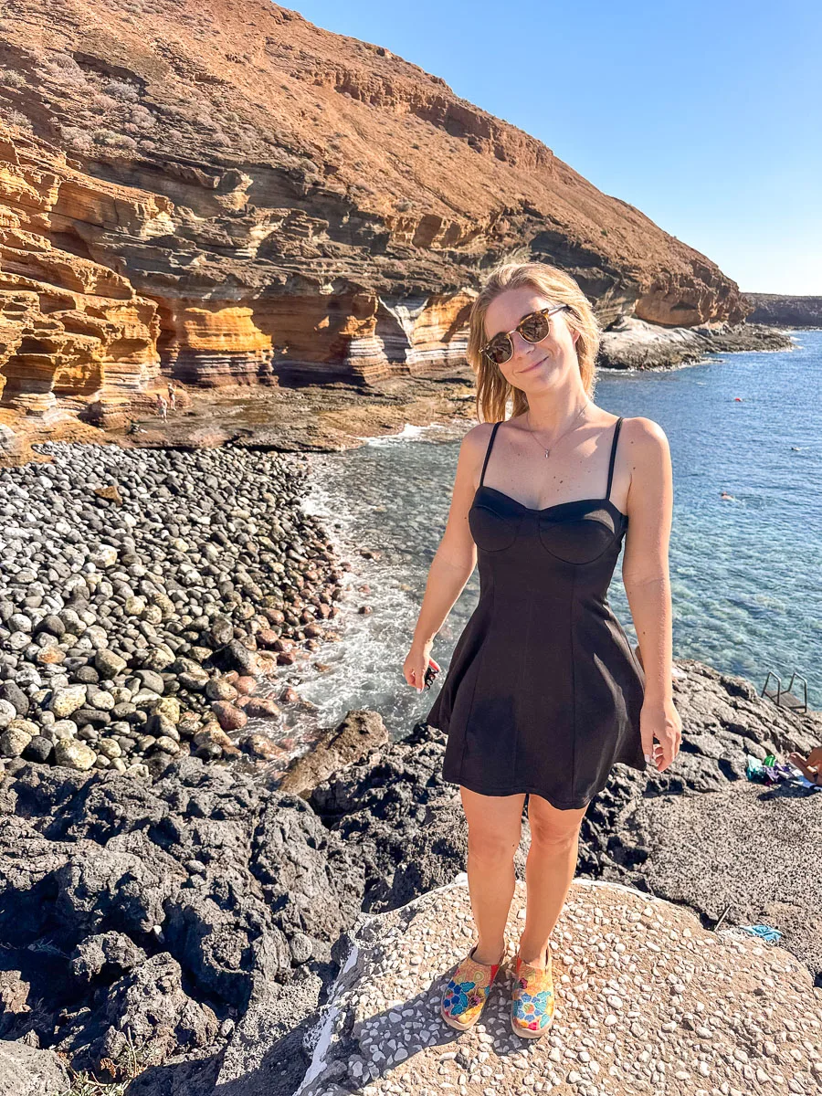 A second view of the author at the same black pebble beach in Tenerife, standing this time, with the sun casting a warm glow on her face. She's wearing a casual black dress and sunglasses, with colorful sneakers that add a playful touch. The calm sea and the cliff's geological layers create a peaceful and scenic setting.