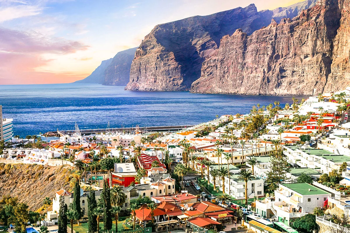A breathtaking view of Los Gigantes, Tenerife, showing the towering cliffs as a backdrop to a quaint coastal town. The white and terracotta-colored buildings contrast with the deep blues of the sea and the sky, while the greenery adds a touch of lushness to the landscape, encapsulating the vibrant life at the edge of the dramatic cliffs.