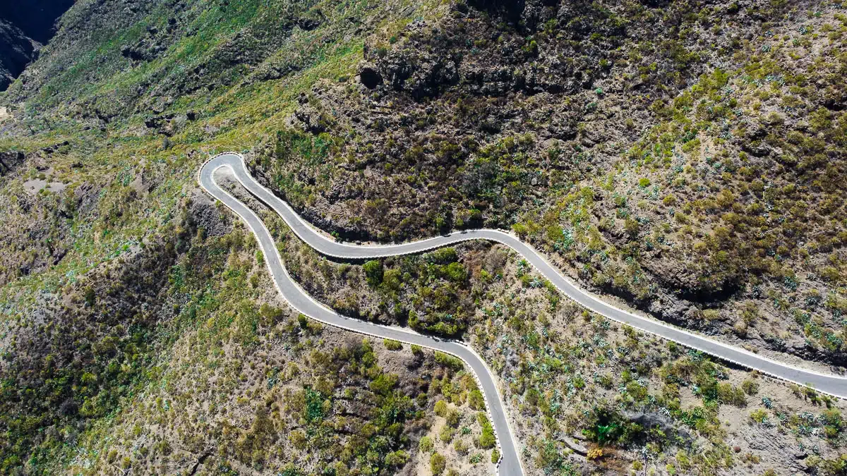 An impressive winding road snakes through the green-covered hills of Tenerife, showcasing the island's diverse terrain and the engineering feat of its roadways. The road cuts a clear path through the otherwise wild and untamed landscape, inviting travelers to explore the hidden corners of the island.