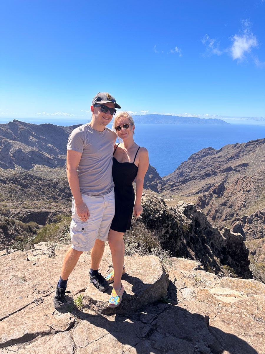 The author and her husband stand atop a rocky peak in Tenerife, with the vast blue sea stretching to the horizon behind them. They are smiling, enjoying the moment, with the clear sky above and the majestic landscape around them. The island of La Gomera is faintly visible in the distance, adding to the sense of adventure and exploration.