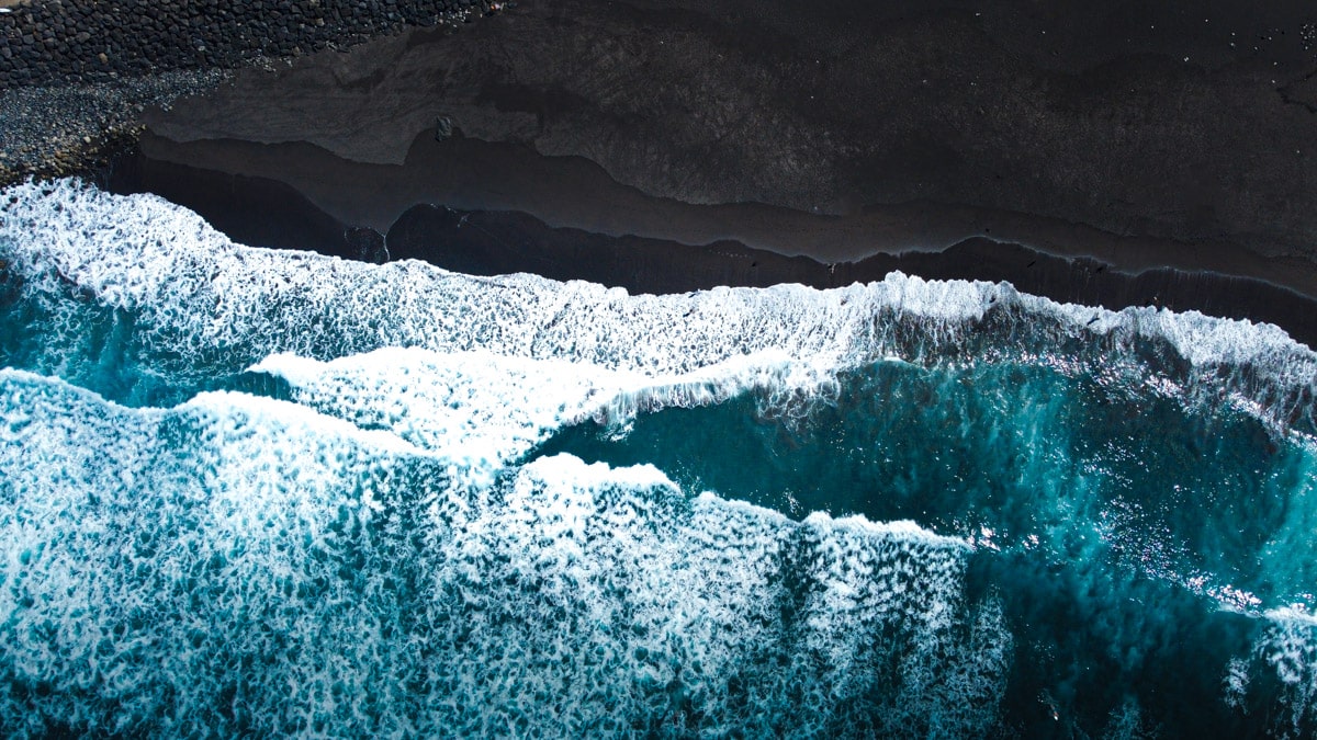 An aerial view of a black sand beach in Tenerife, where the powerful Atlantic waves create frothy white patterns as they break onto the shore, contrasting with the dark volcanic sand and the parallel parked cars along the coastal road.