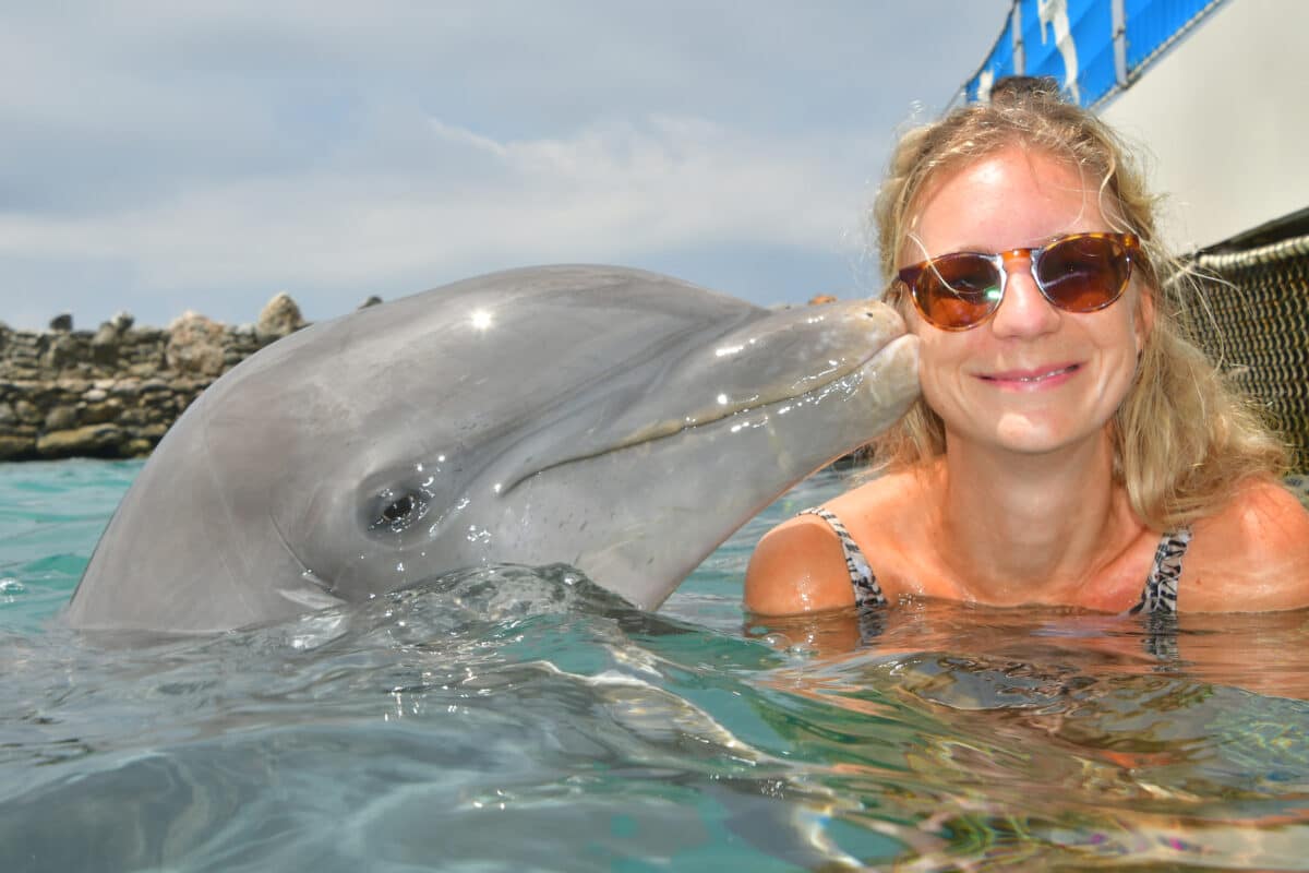 A woman in a zebra-print swimsuit gives a peace sign while interacting with a playful dolphin in the clear waters of Curaçao, an unforgettable experience for marine life enthusiasts. you can see the author sabrina in the picture