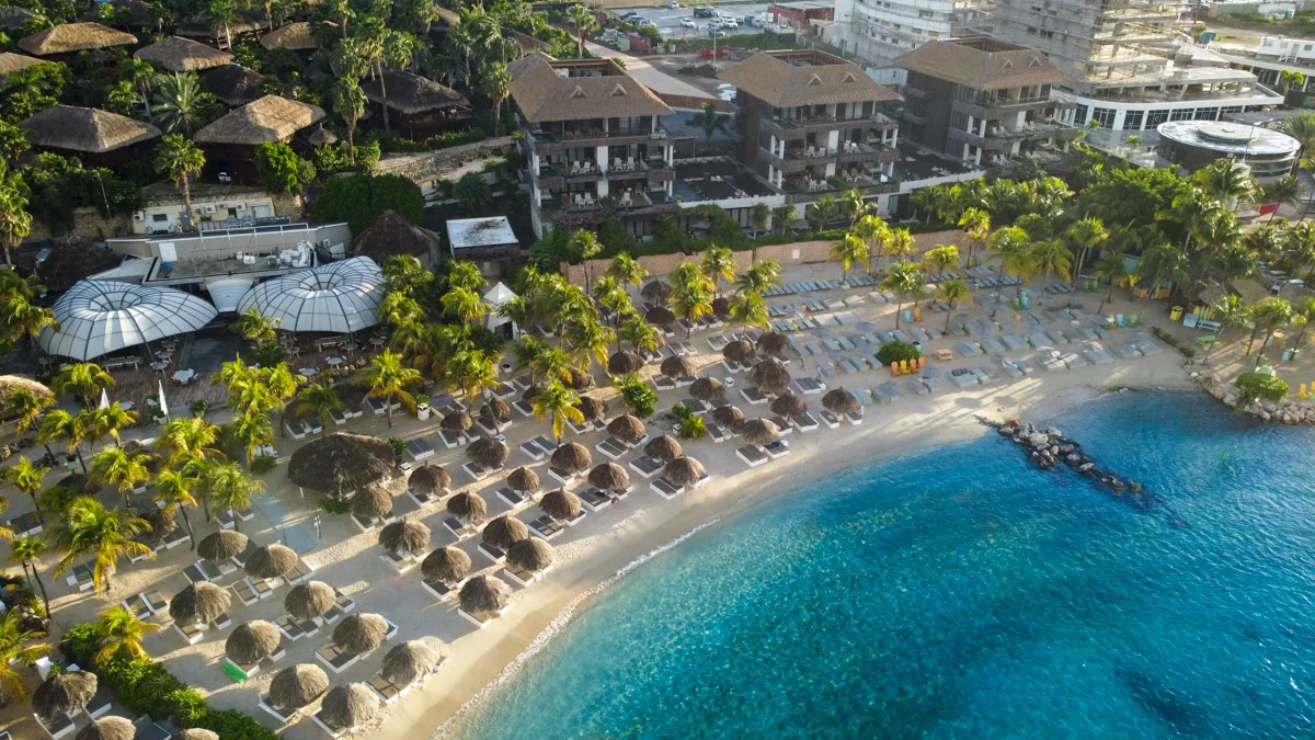 An aerial shot capturing the serene beauty of a Curacao dive resort, showcasing thatched umbrellas on a pristine sandy beach with crystal-clear turquoise waters inviting exploration.