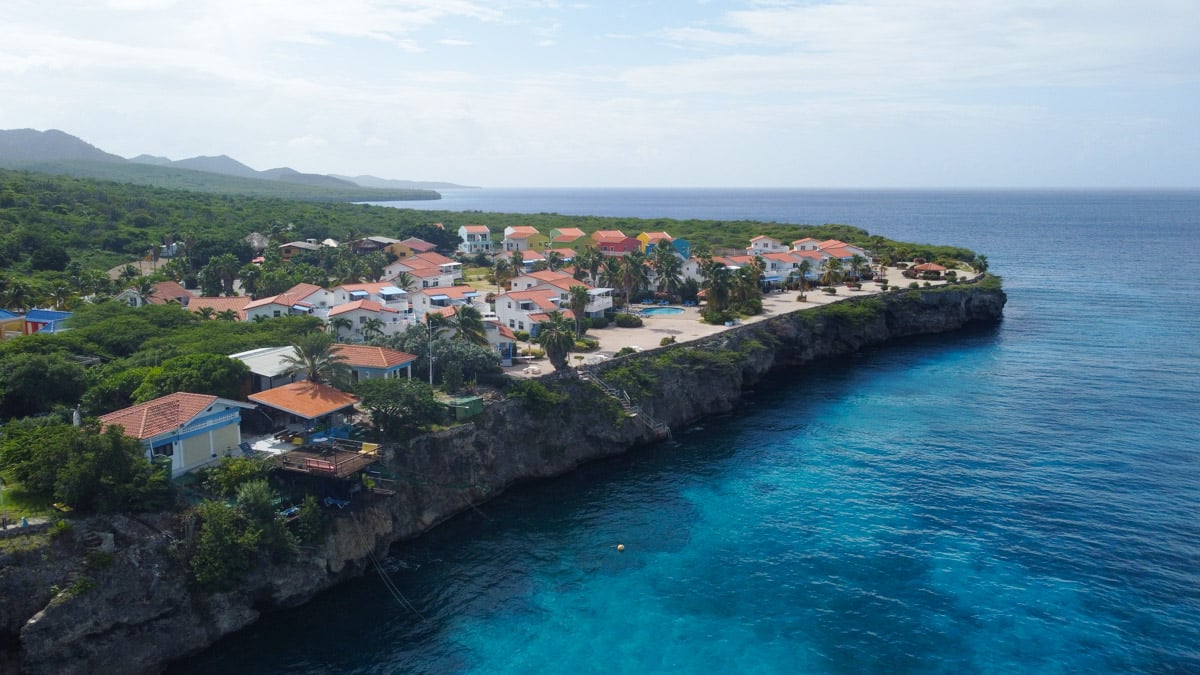 An expansive aerial view of a tranquil residential area in Curacao, perched on a cliff overlooking the azure sea, hinting at the hidden dive spots nearby. You can see the cute houses of the Marazul resort in this picture