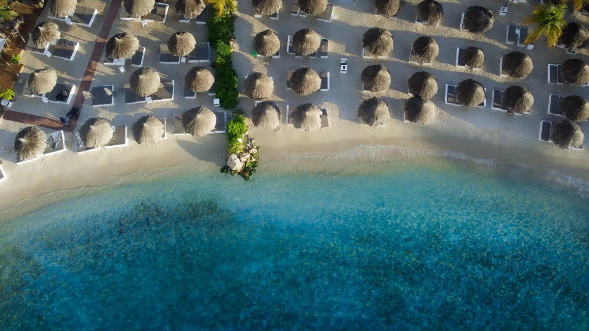 Aerial view of Mambo Beach in Curaçao, showcasing a resort with thatched umbrellas and sun loungers on a sun-kissed sandy beach with clear turquoise waters.