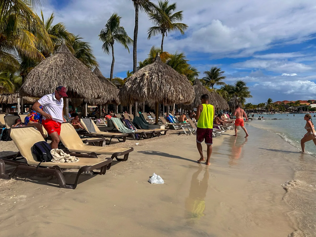 A lively beach scene at Mambo Beach in Curaçao, featuring vacationers and a bright yellow lifeguard, with thatched umbrellas and palm trees swaying in the breeze.
