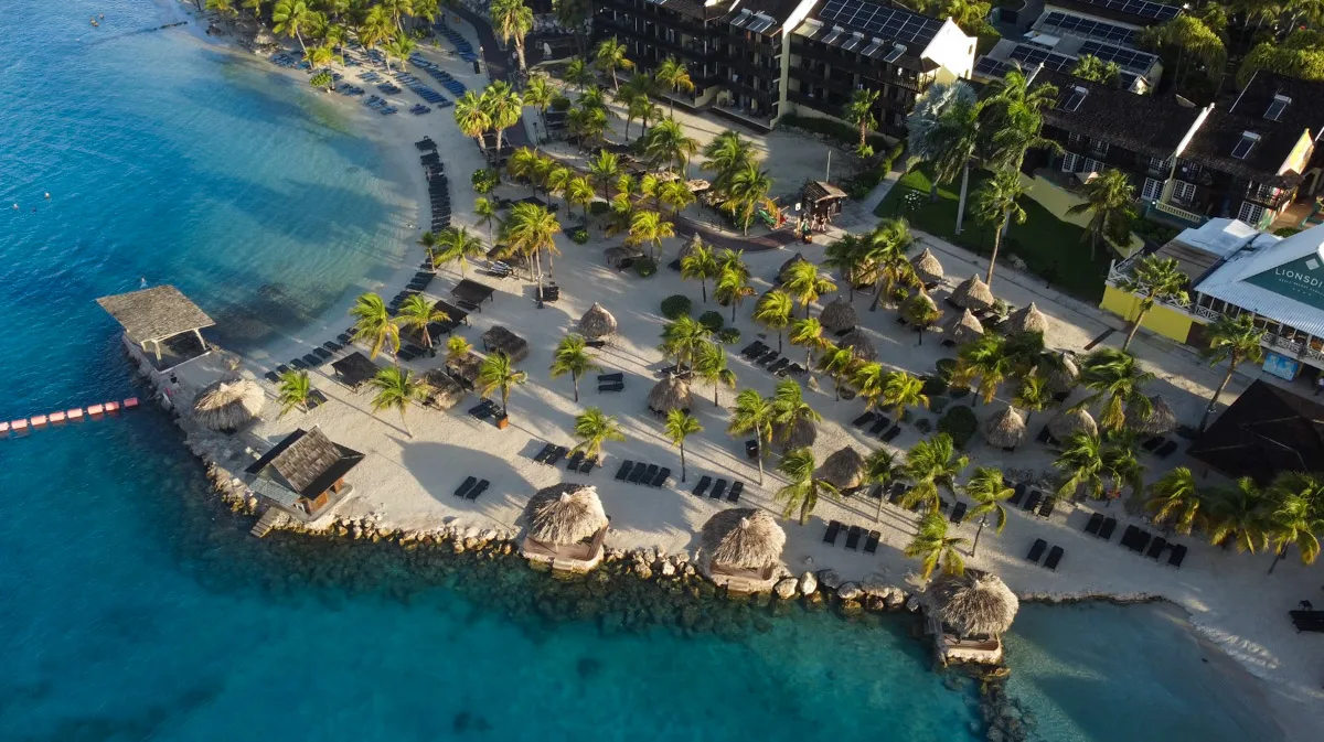 A sweeping aerial view of the bustling Mambo Beach in Curaçao, with beachfront resort Lions Dive nestled among greenery and the island's signature clear blue waters.